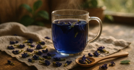 A glass mug of vibrant blue butterfly pea flower tea sits on a linen cloth, surrounded by dried butterfly pea blossoms and a wooden spoon